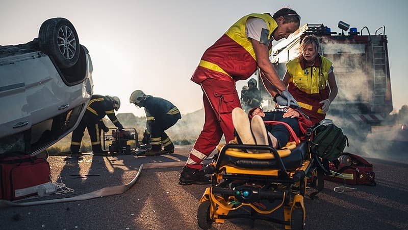 Paramedics and firefighters respond to a serious car accident, stabilizing an injured person on a stretcher while others manage equipment near an overturned vehicle.